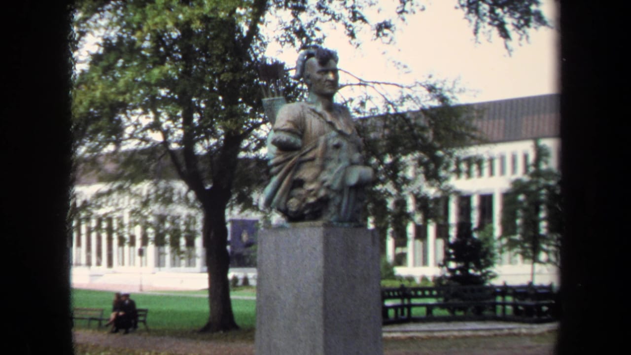 1970 Annapolis home-movie frame of a Native American warrior statue on a stone pedestal, with benches, trees, lawn, and a distant seated couple.