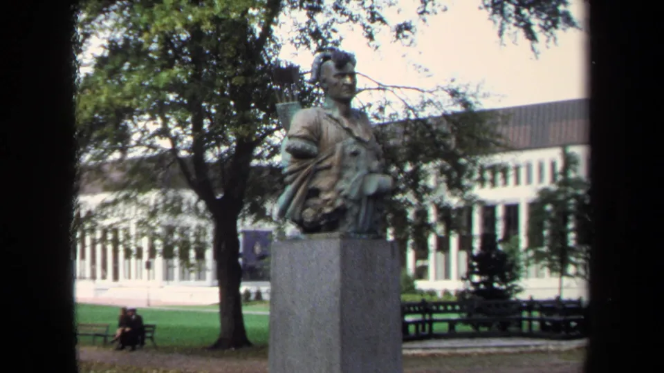 1970 Annapolis home-movie frame of a Native American warrior statue on a stone pedestal, with benches, trees, lawn, and a distant seated couple.