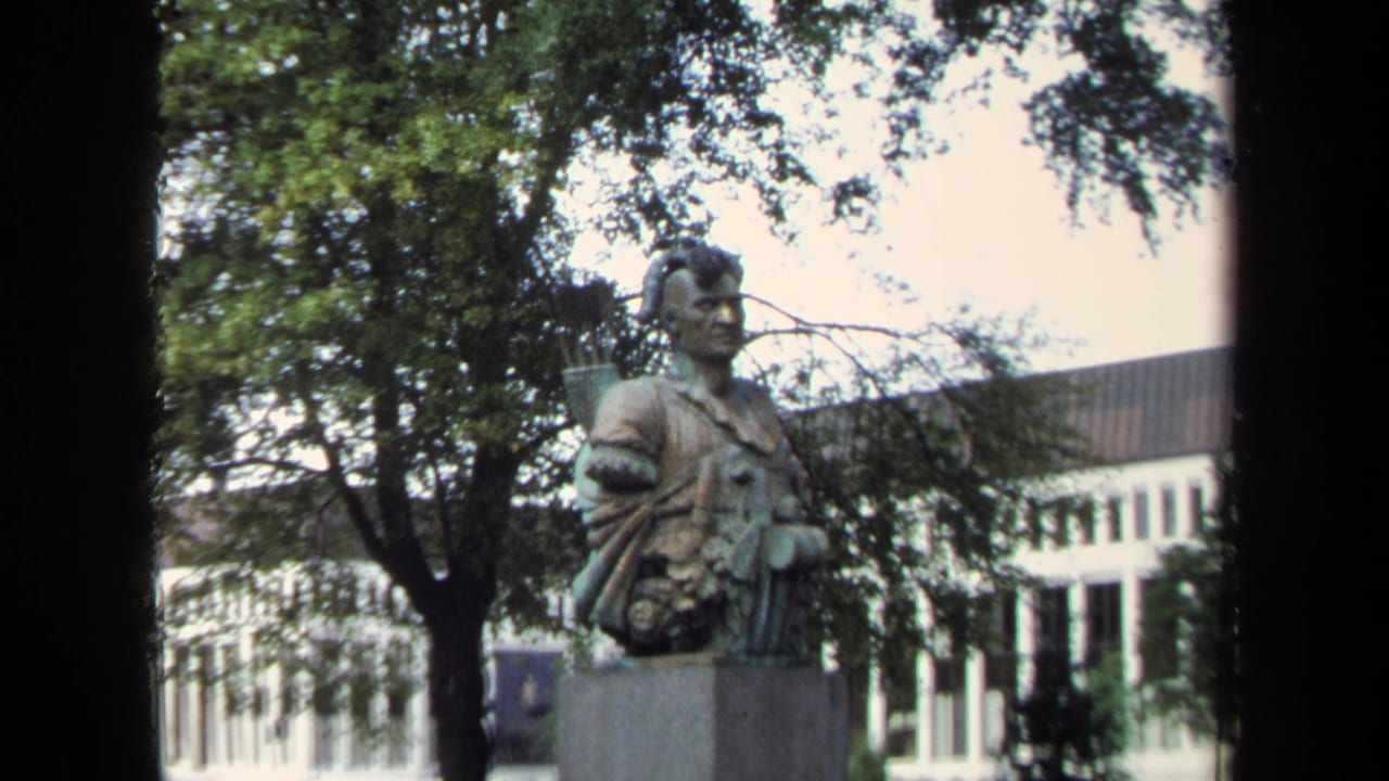 1970 Annapolis frame showing the warrior bust and arrow quiver from a slightly closer angle, partly framed by trees and the white building.