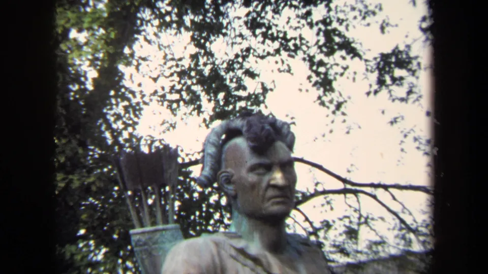Tight 1970 Annapolis frame of the bronze-toned warrior statue's head and upper body, with arrows and dense tree leaves in the background.