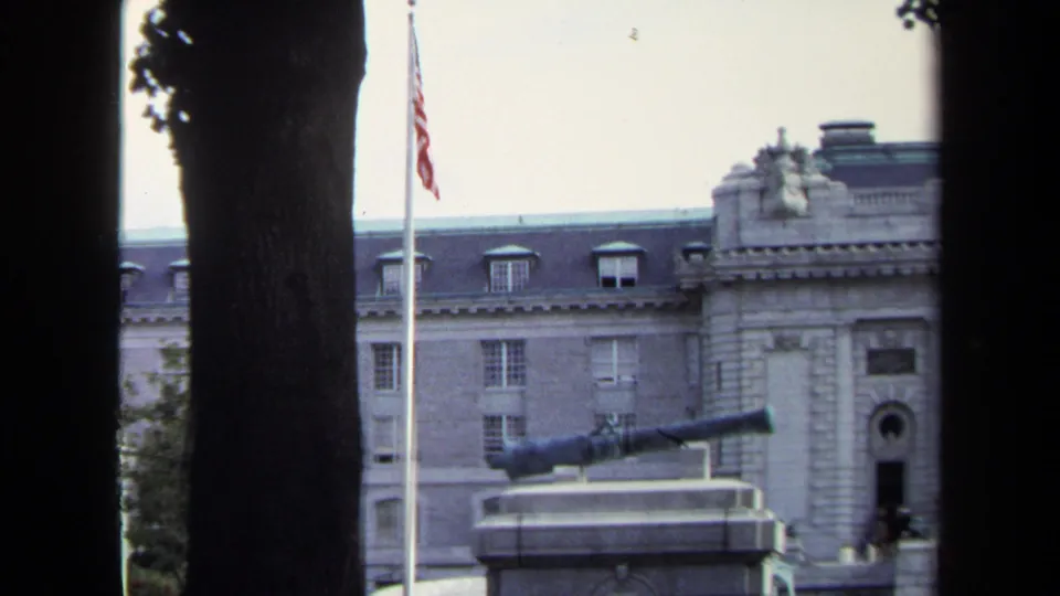 1970 Annapolis frame with a U.S. flag, cannon display, tree trunk, and stone building exterior.