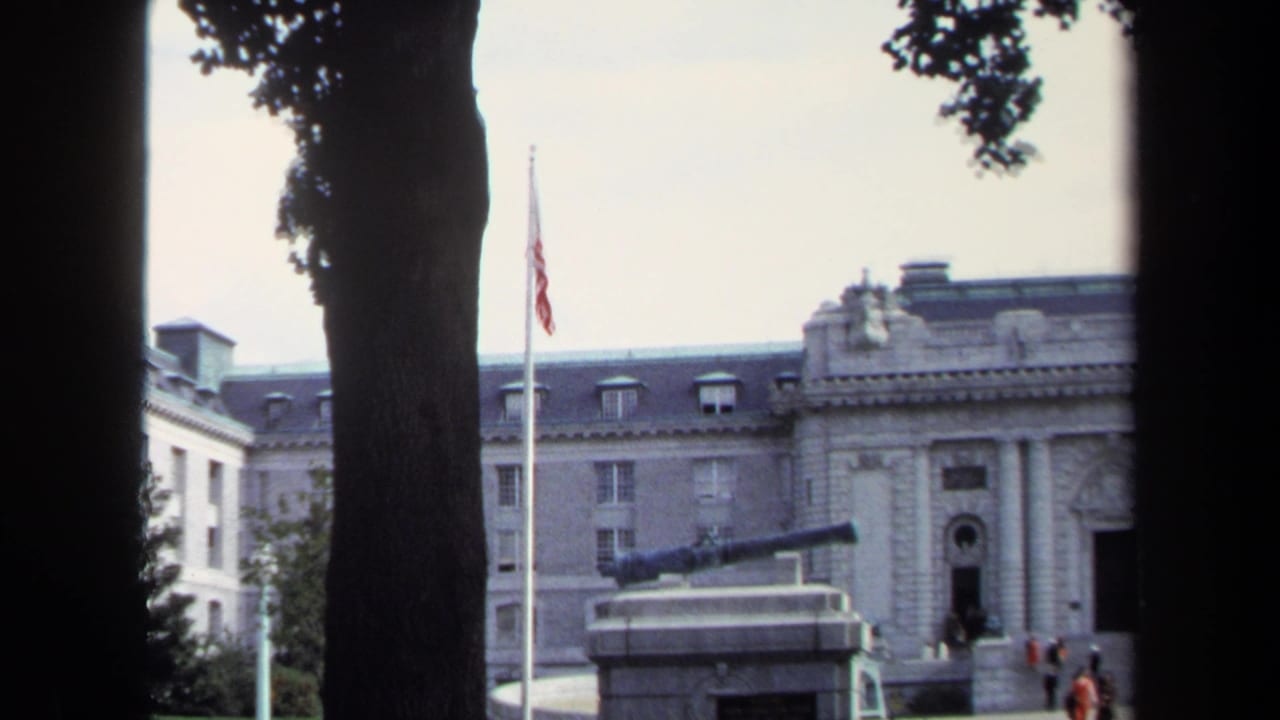 1970 Annapolis home-movie frame showing a formal stone government building, half-mast American flag, tree trunks, and a cannon monument.