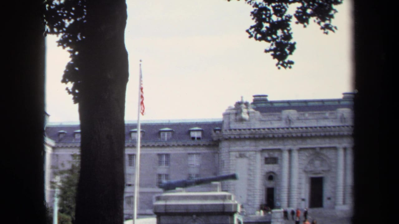 Cannon monument and flagpole in front of a large stone building, partially framed by trees.