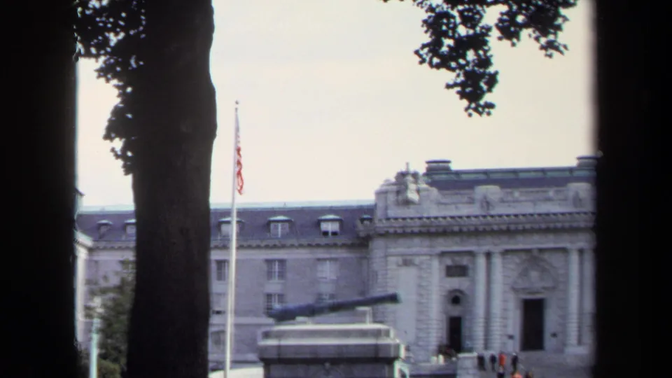 1970 Annapolis frame with a cannon monument, half-mast American flag, stone civic facade, and small visitors near the entrance.