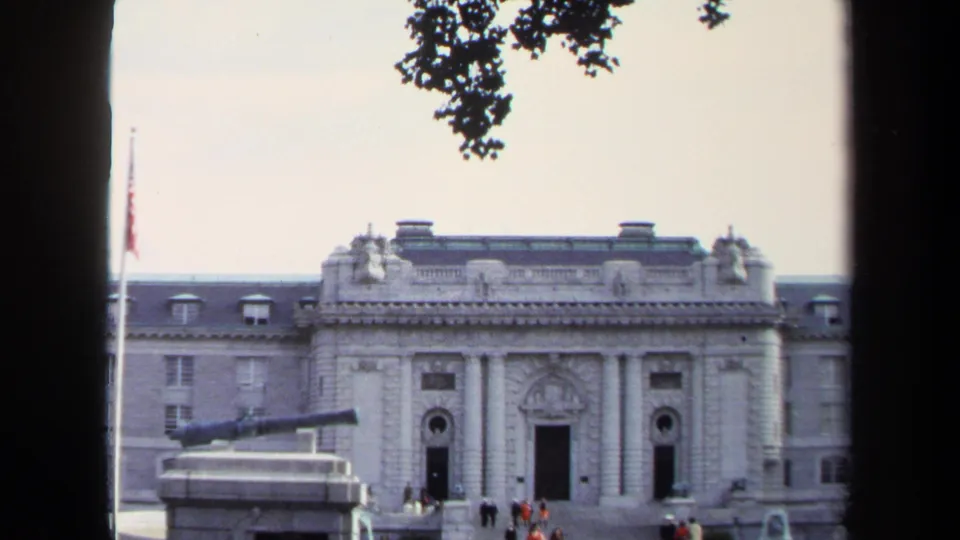1970 Annapolis home-movie view of visitors outside a formal stone government building with columns, flagpole, and cannon monument.
