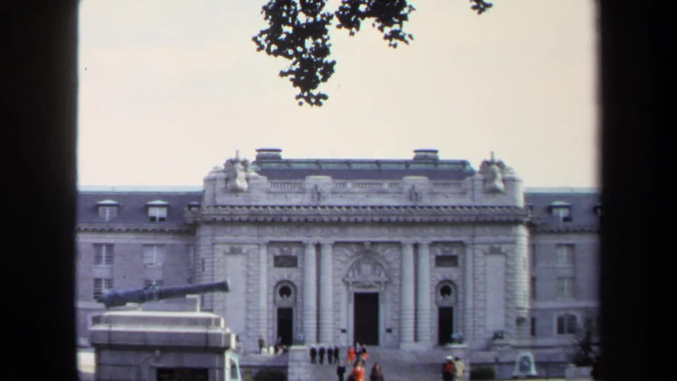 1970 Annapolis frame showing a stone civic building, cannon monument, shaded trees, and visitors gathered near the entrance steps.
