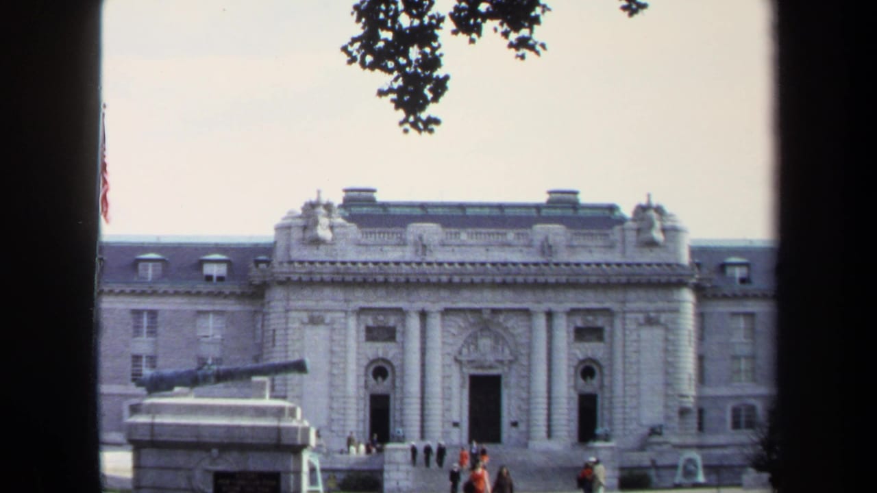 1970 Annapolis home-movie frame showing a formal stone building, visitors near the entrance, a small cannon at left, and dark film-frame edges.
