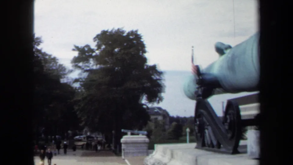 1970 Annapolis home-movie view of the green cannon from the side, with trees, an American flag, stonework, and distant visitors visible.