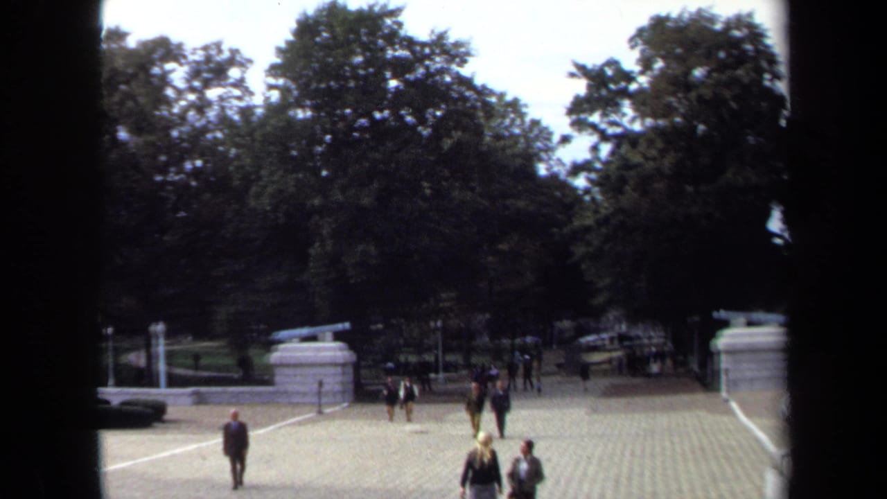 1970 Annapolis home-movie frame showing formally dressed adults crossing a sunlit courtyard near trees and stone cannon mounts.