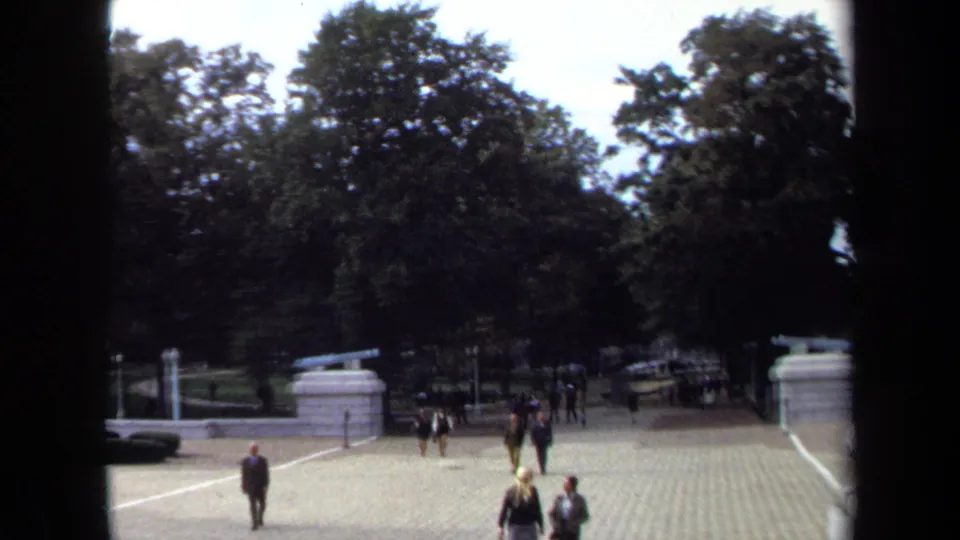 1970 Annapolis home-movie frame showing formally dressed adults crossing a sunlit courtyard near trees and stone cannon mounts.