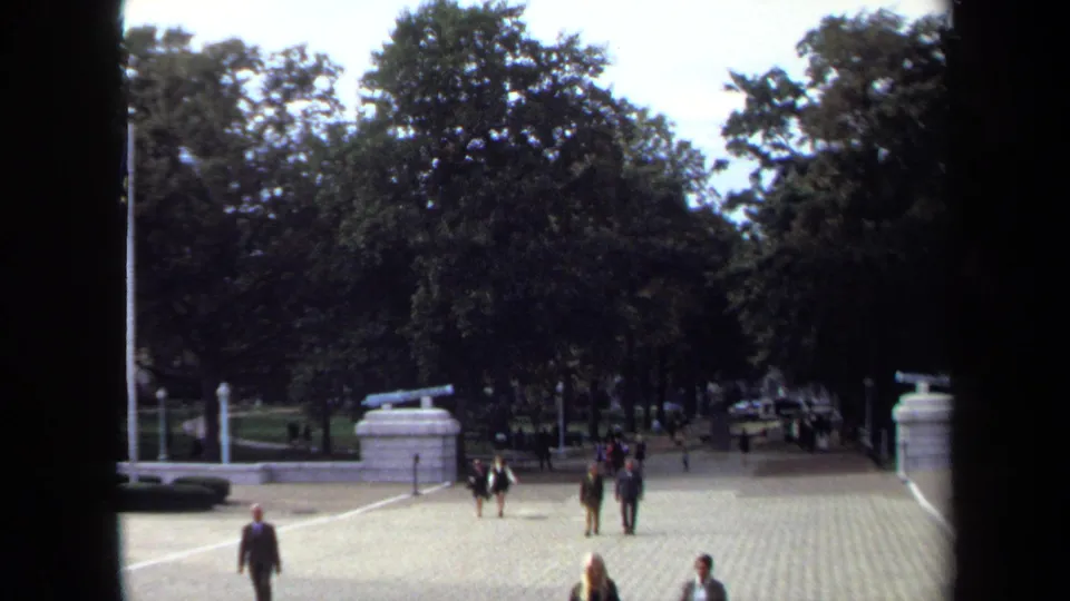 1970 Annapolis home-movie view of adults crossing a broad paved courtyard with mature trees and stone cannon mounts nearby.