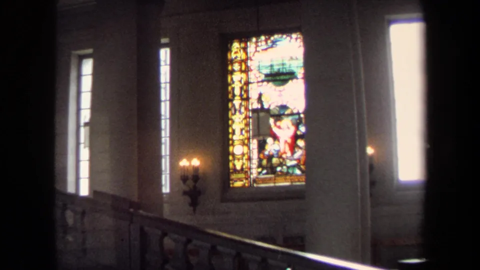 1970 Annapolis interior frame showing a glowing stained-glass religious window, tall windows, wall sconces, and an upper railing.