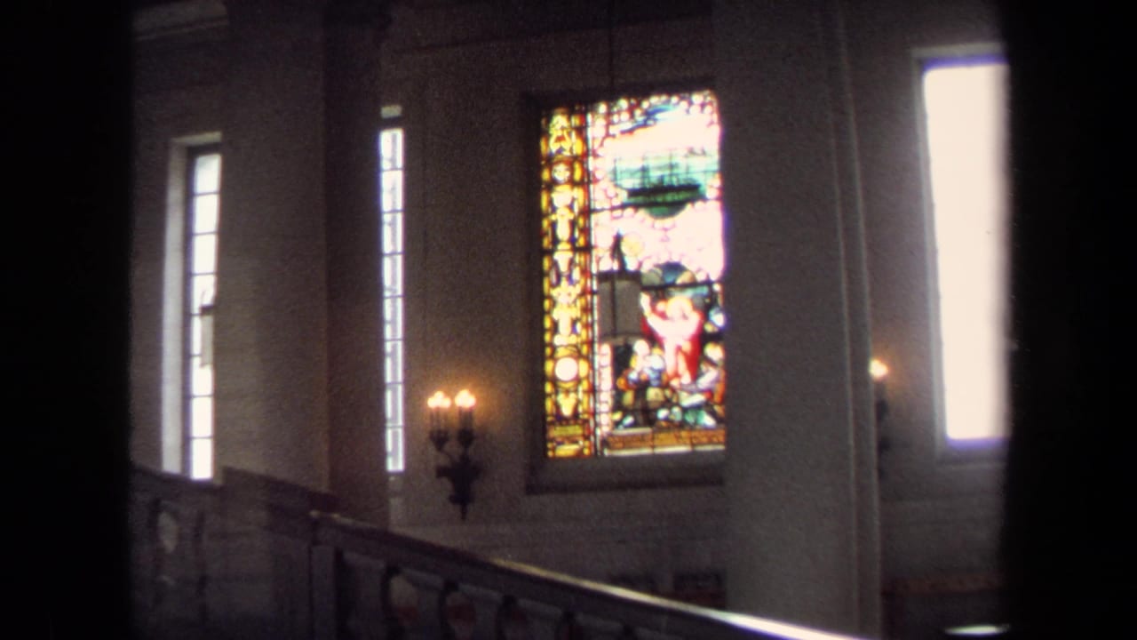 1970 Annapolis home-movie frame showing a dim church interior with stained glass panels, tall windows, candle sconces, and foreground railing.
