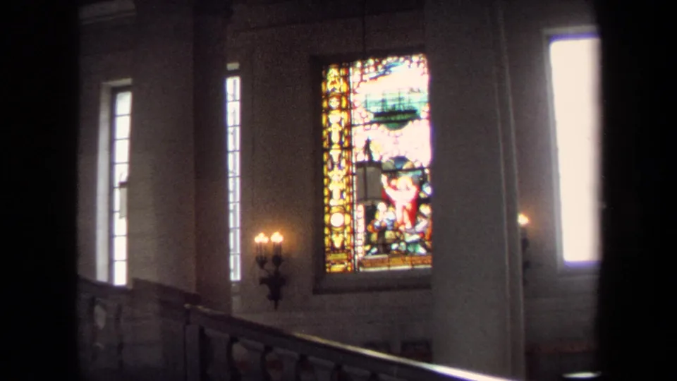 1970 Annapolis home-movie frame showing a dim church interior with stained glass panels, tall windows, candle sconces, and foreground railing.