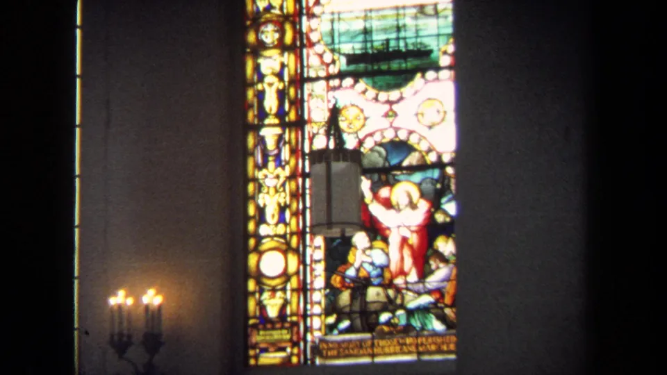 1970 Annapolis home-movie close view of a stained glass panel with religious figures, a ship motif, and warm candlelight below.