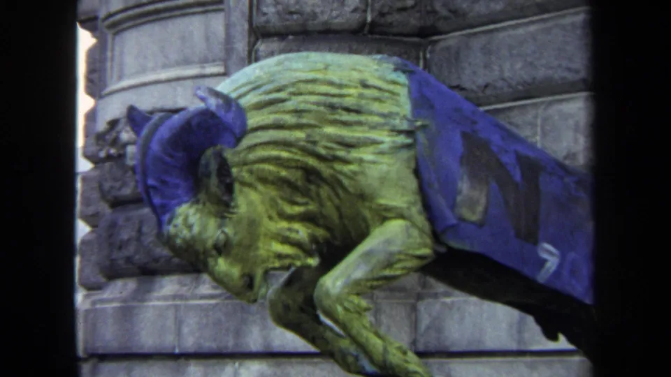 Close 1970 Annapolis home-movie view of a weathered ram or goat-like sculpture draped in blue fabric near stone architecture.