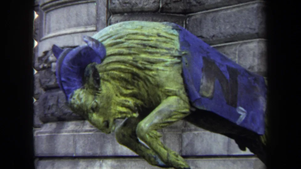 1970 Annapolis frame of a mascot-like animal sculpture wearing blue cloth with a large N, set against a stone building facade.