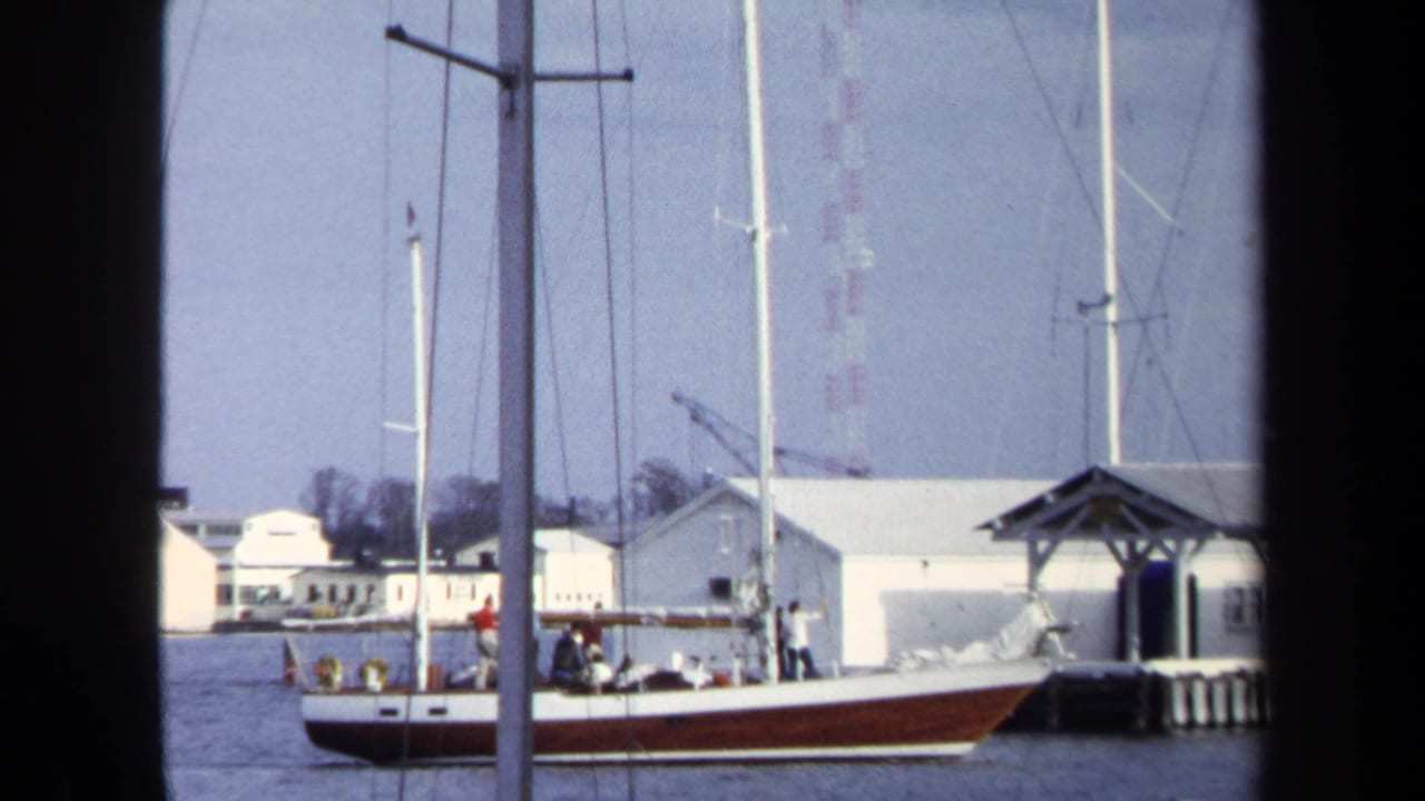 Red-and-white sailboat near a dock with tall masts and white buildings behind it.