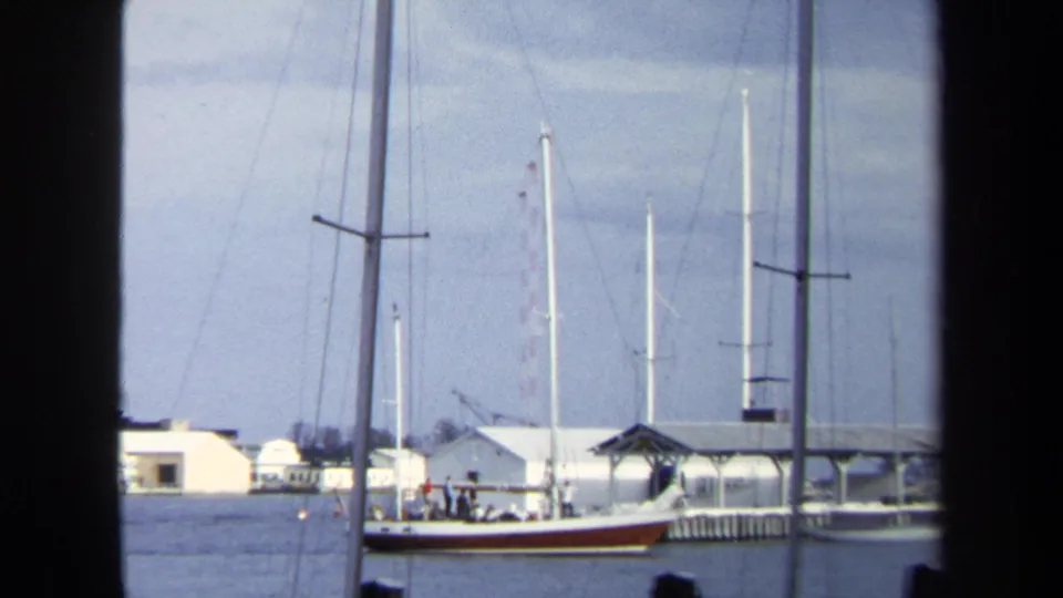 The sailboat and marina buildings appear through dark vertical window framing, with masts, water, dock structures, and distant people visible.