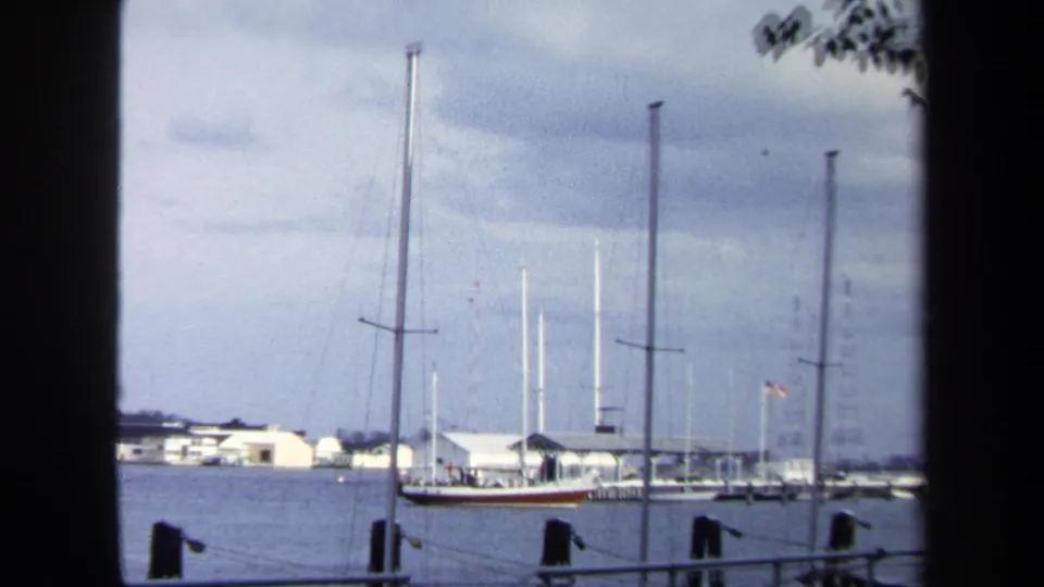 A wider Annapolis harbor view shows the sailboat near docks, multiple masts, low white buildings, water, and a gray-blue cloudy sky.