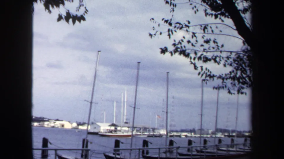 Annapolis waterfront marina view from 1970 8mm footage, with sailboats moored beside dock posts and trees edging the frame.
