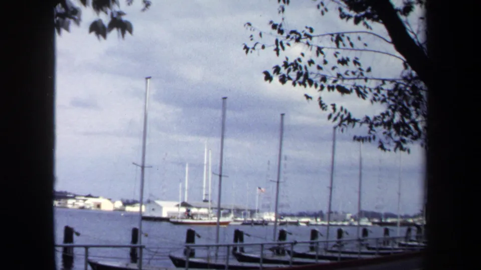 1970 Annapolis 8mm harbor frame showing sailboats and masts across calm water, with dock rails and leafy branches in view.