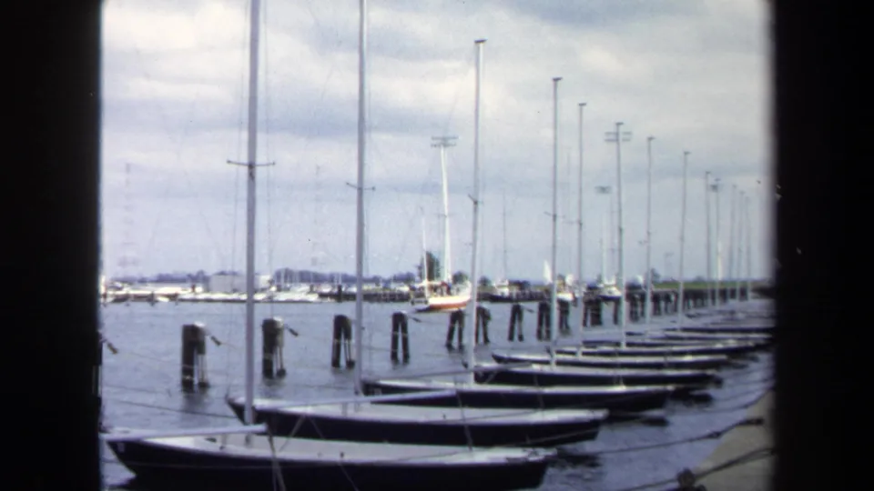 1970 Annapolis marina frame with small boats pulled along the dock, sailboat masts, wooden pilings, calm water, and a cloudy sky.
