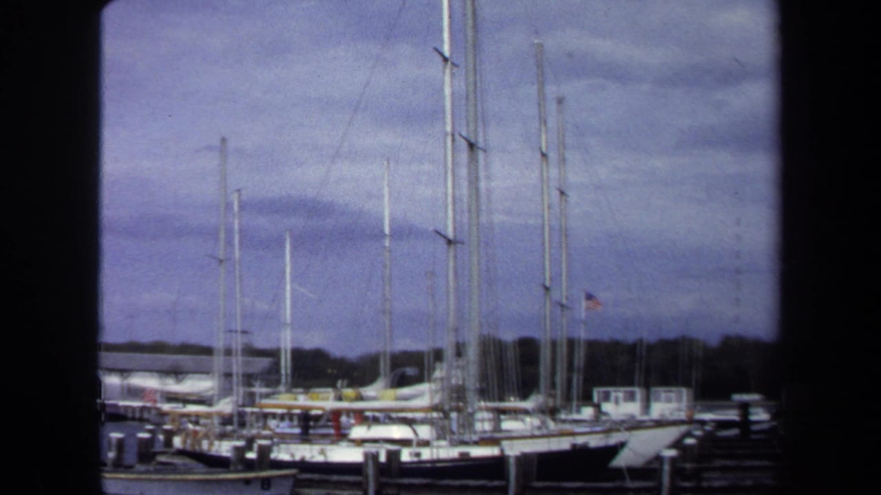 1970 Annapolis marina view with docked sailboats, tall masts, wooden piers, cloudy sky, and a dark frame in the foreground.
