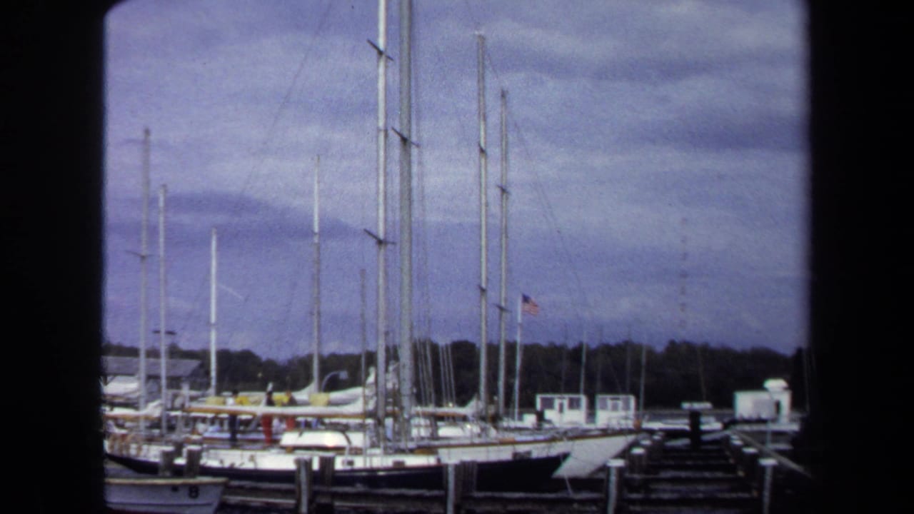 1970 Annapolis sailboats sit moored beside wooden piers, with a dark foreground divider and overcast sky above the masts.