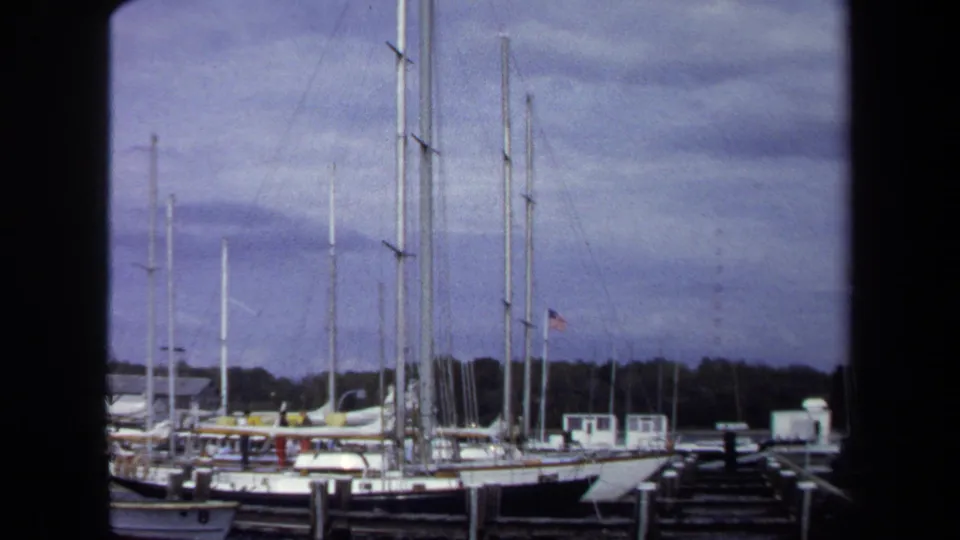 1970 Annapolis sailboats sit moored beside wooden piers, with a dark foreground divider and overcast sky above the masts.