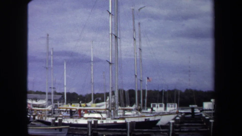 1970 Annapolis marina scene showing clustered sailboat masts, docked boats, piers, cloudy sky, and a dark home-movie frame edge.