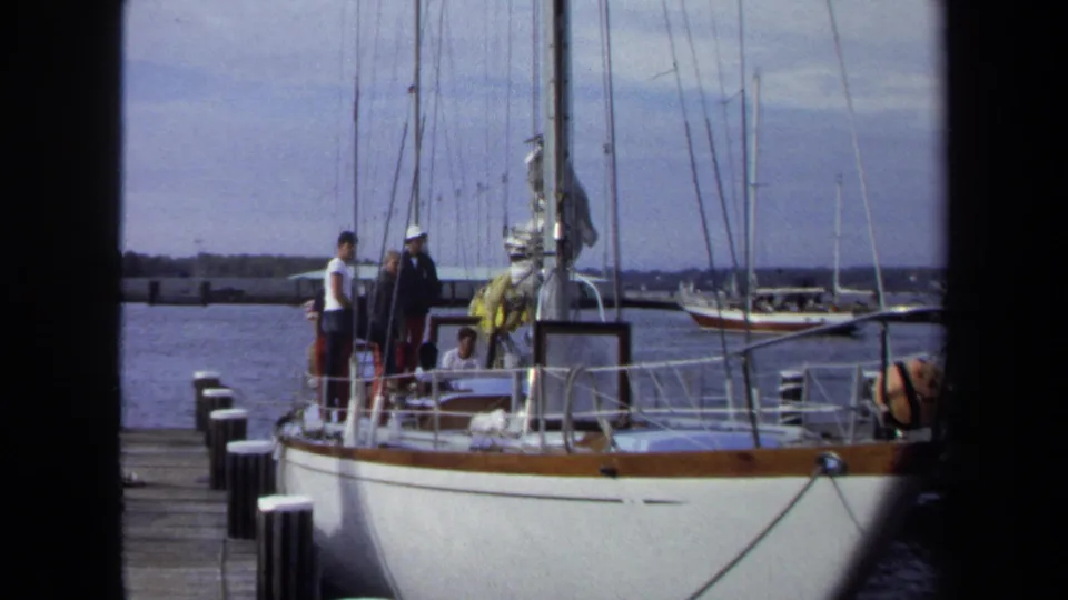 1970 Annapolis marina footage shows people in casual clothing aboard a white sailboat near a wooden dock, with masts and water behind them.
