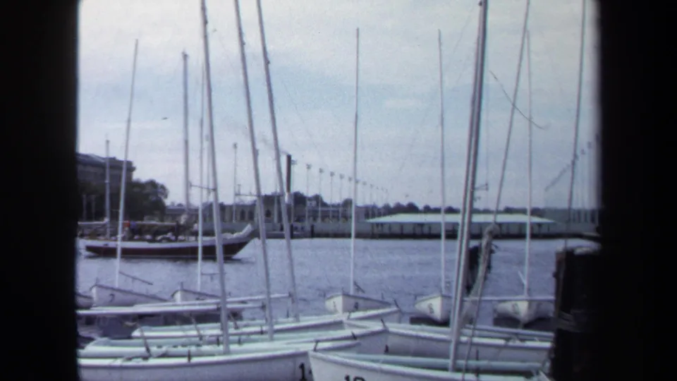 1970 Annapolis marina view with small docked boats in front, a larger sailboat beyond, and low buildings under a cloudy sky.