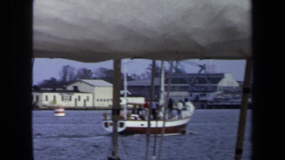 Annapolis harbor footage from 1970 shows a sailboat carrying several distant people moving past pier posts and low waterfront buildings.