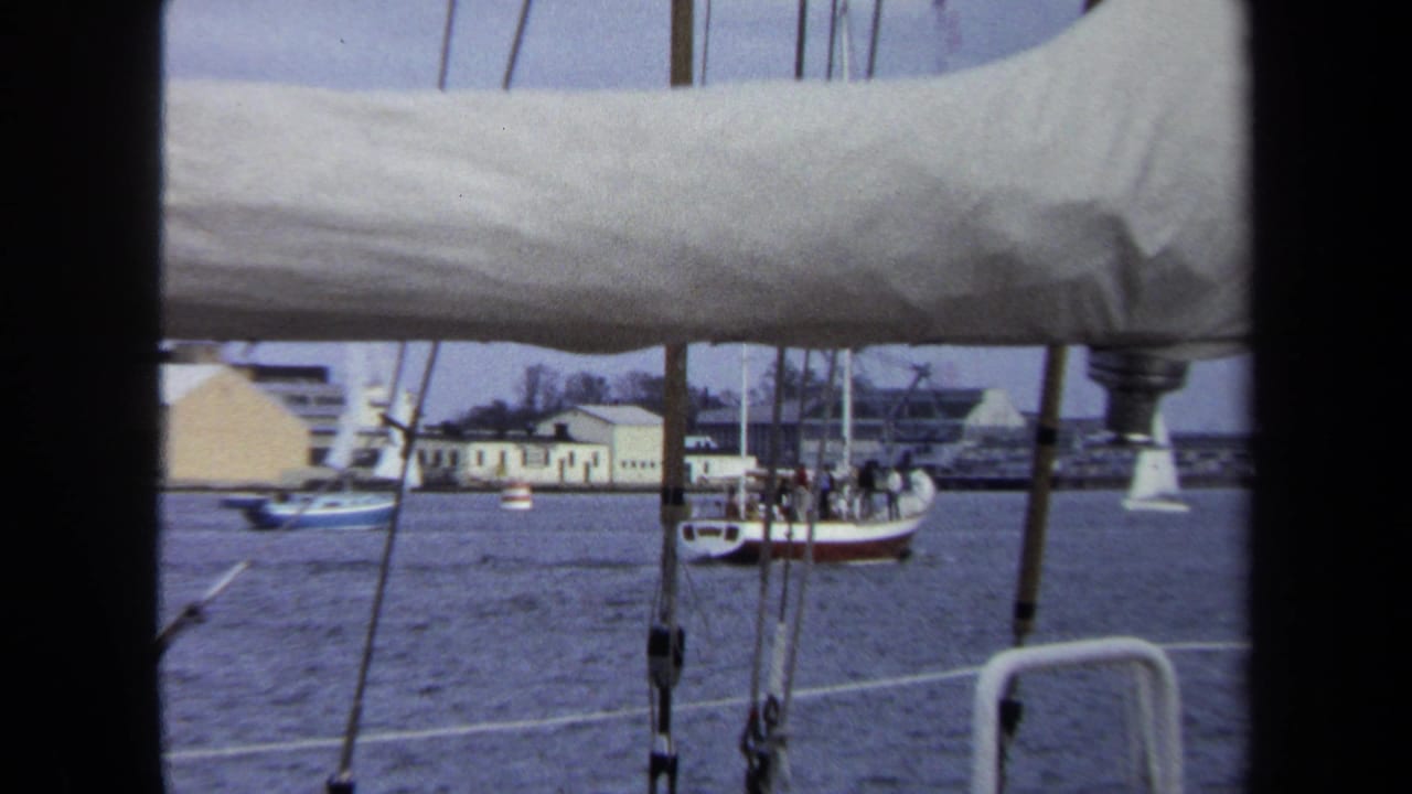 1970 8mm Annapolis view from a sailboat deck, with rigging, a furled sail, water, distant buildings, and a small boat carrying people.