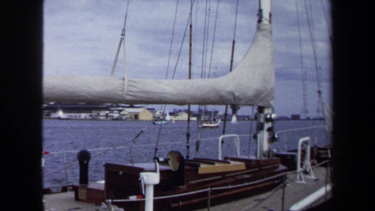 Wood-trimmed sailboat deck and railings with water, rigging, and distant shore buildings beyond.