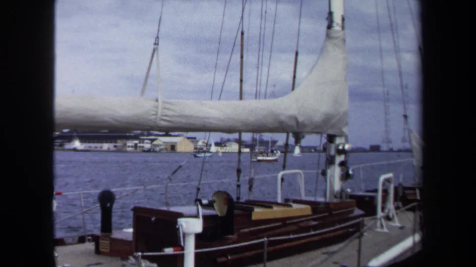 Annapolis sailboat deck scene with varnished wood, white railings, furled sail, rigging, blue water, and distant waterfront structures.