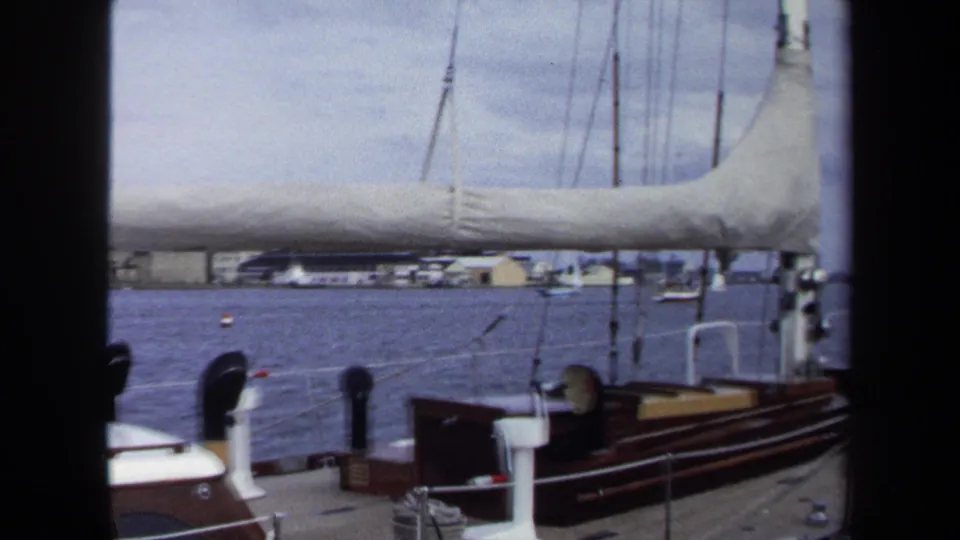 1970 home-movie frame from an Annapolis sailboat, showing deck fittings, rigging, furled sail, harbor water, and low buildings ashore.