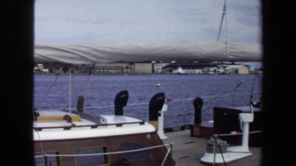 Annapolis 8mm frame showing the sailboat cabin top and railings under a furled sail, with blue water and low shore buildings beyond.