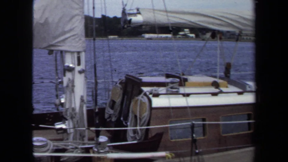1970 Annapolis home-movie frame with a mast, sail cover, cabin roof, deck lines, and harbor water.