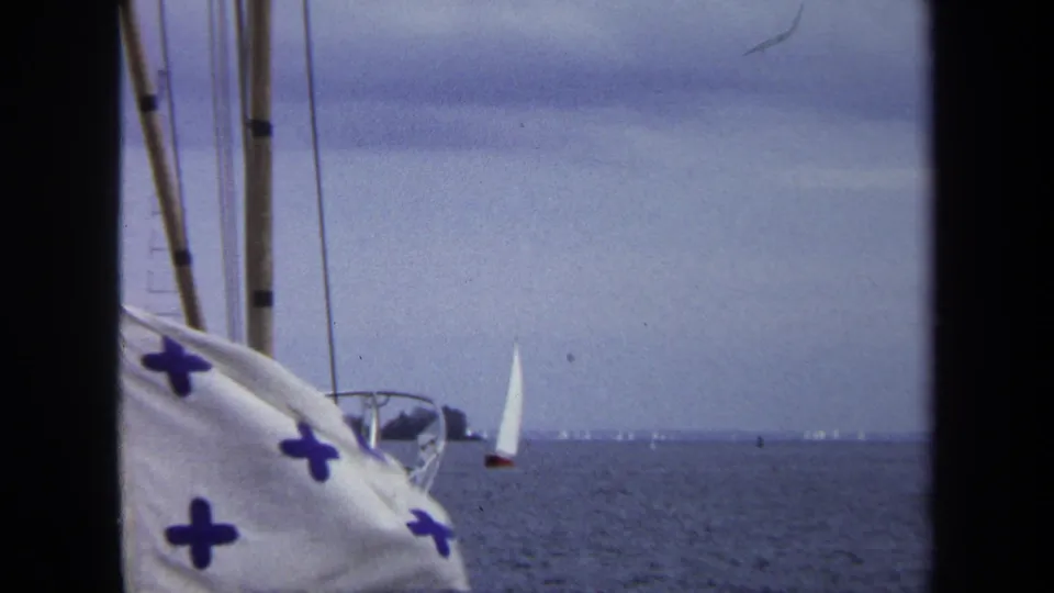 1970 Annapolis home-movie frame with rigging, a white flag with blue crosses, and several distant sailboats on blue water.
