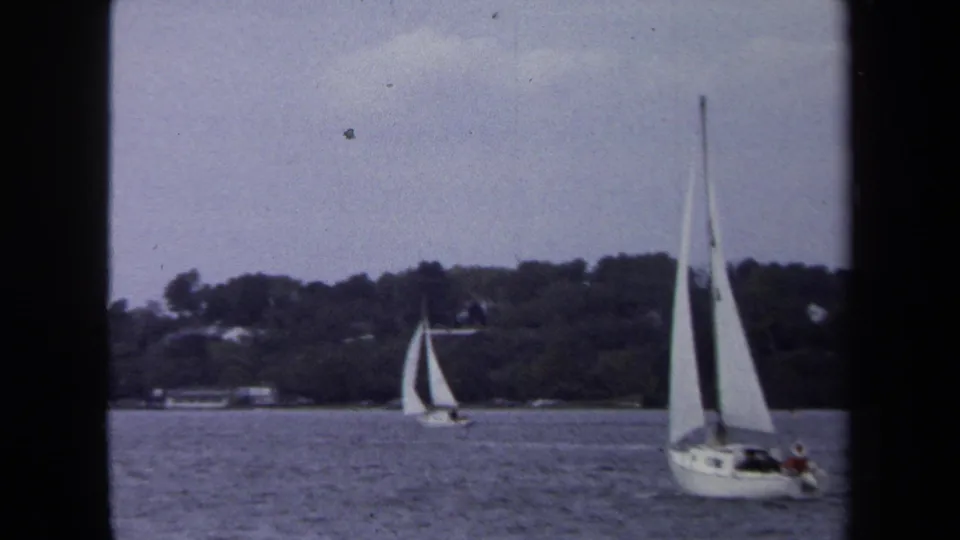 1970 Annapolis 8mm frame of sailboats near a wooded shoreline, with a larger white sailboat crossing the foreground water.