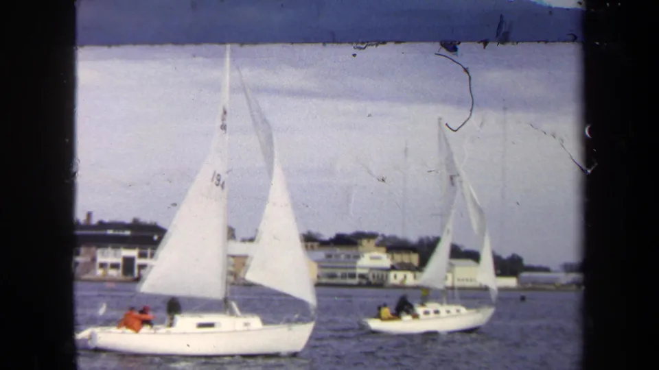 1970 Annapolis-area 8mm frame showing two sailboats with visible occupants near low buildings along the water.