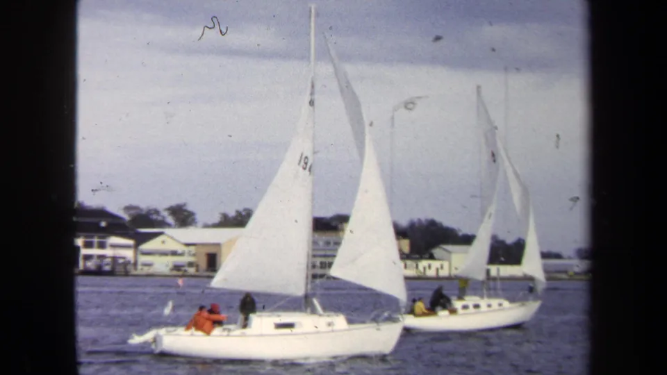 1970 Annapolis-area sailing frame with two small boats, white sails, distant occupants, and low waterfront buildings.