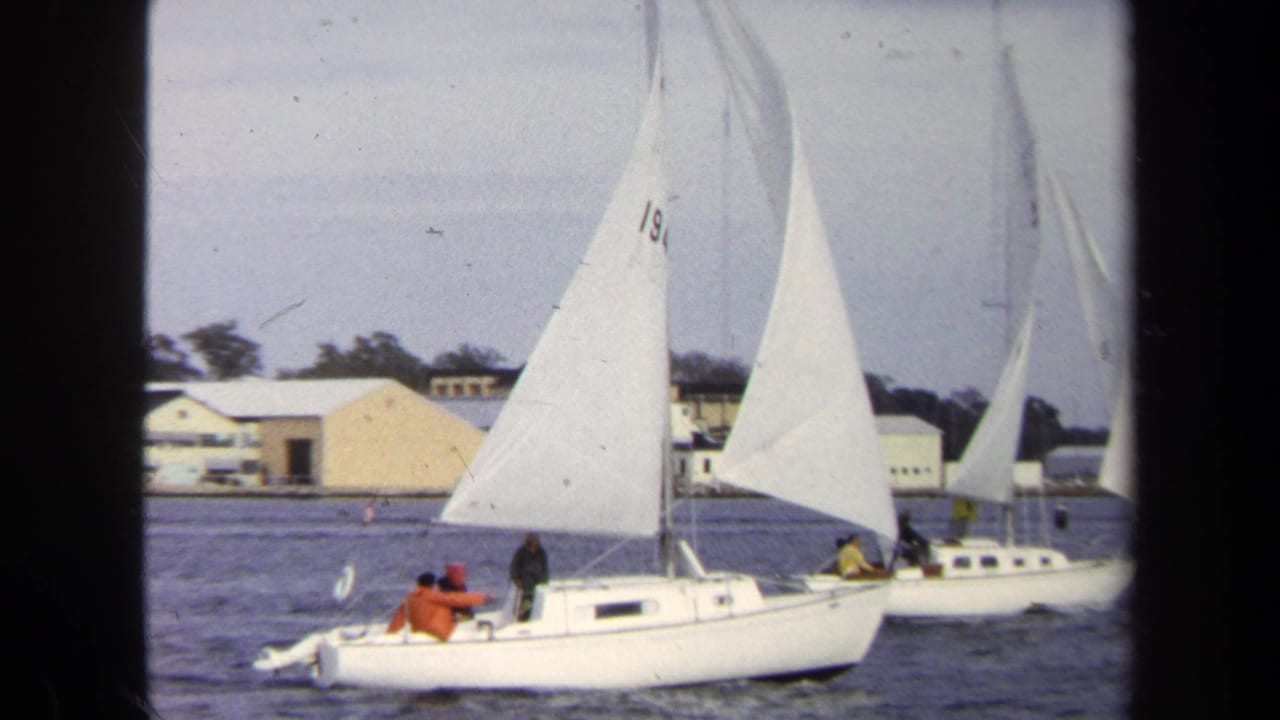 1970 Annapolis home-movie frame showing a white sailboat with crew in bright jackets passing near waterfront buildings.