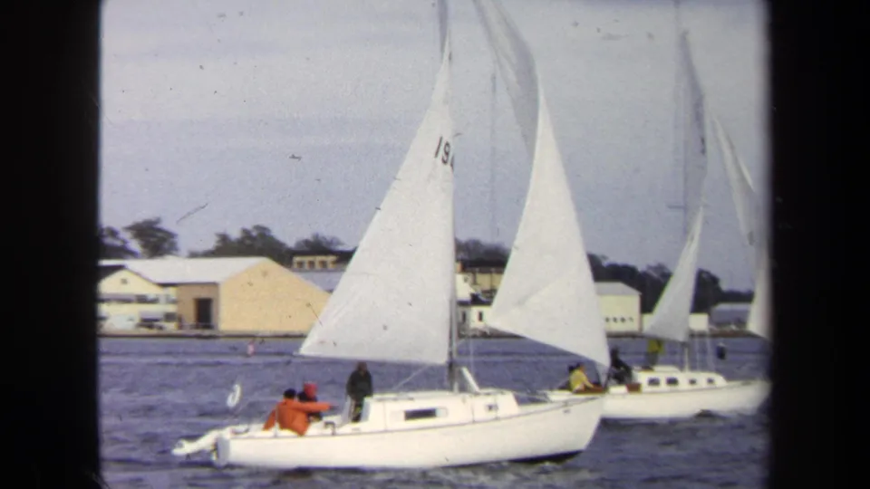 1970 Annapolis home-movie frame showing a white sailboat with crew in bright jackets passing near waterfront buildings.