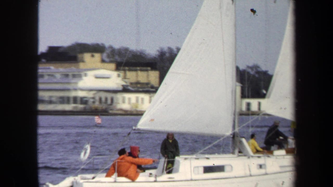 Close view of a white sailboat with people on deck and shoreline behind it.
