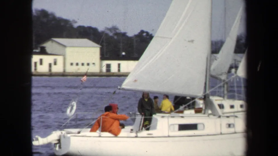 1970 Annapolis 8mm frame showing a close pass of the sailboat, with crew in bright outerwear near the cockpit.