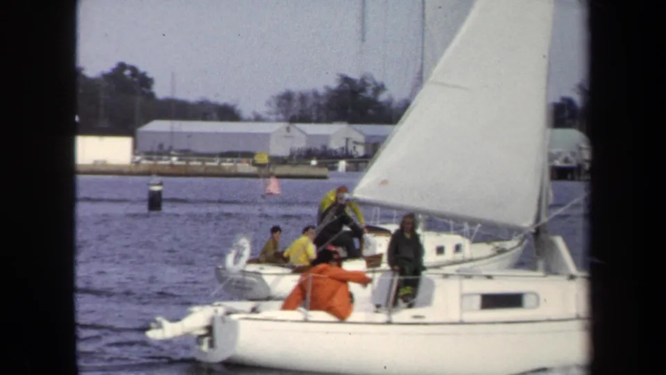 1970 Annapolis home-movie frame showing several people aboard a white sailboat under sail, with buildings and trees across the water.