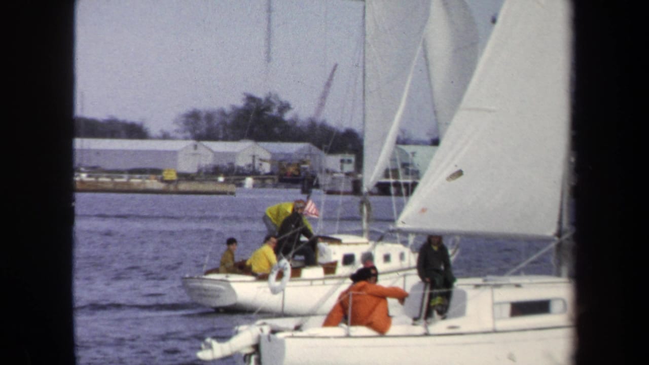 1970 Annapolis home-movie frame of white sailboats with people aboard, seen from another boat near a low waterfront shoreline.